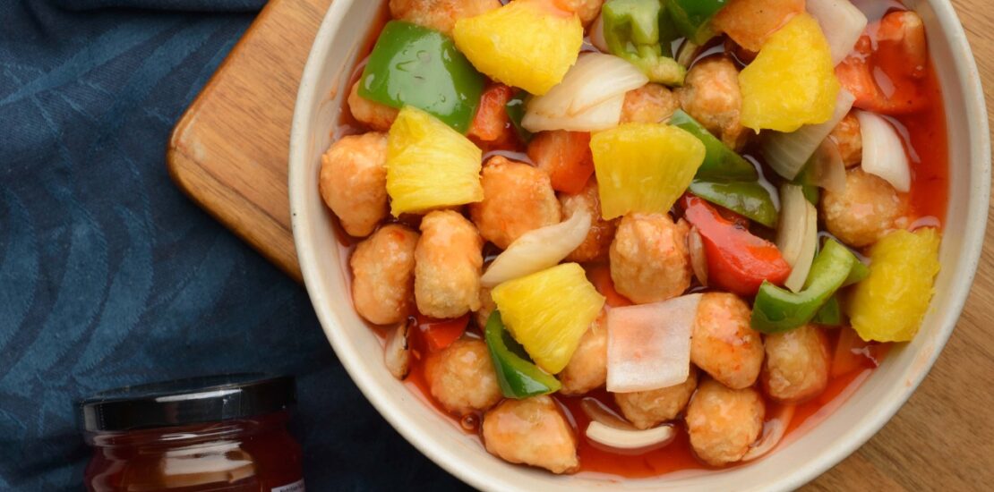 A bowl of sweet and sour chicken with pineapple, bell peppers, and onions sits on a wooden board next to a jar of MF Food sweet and sour sauce.